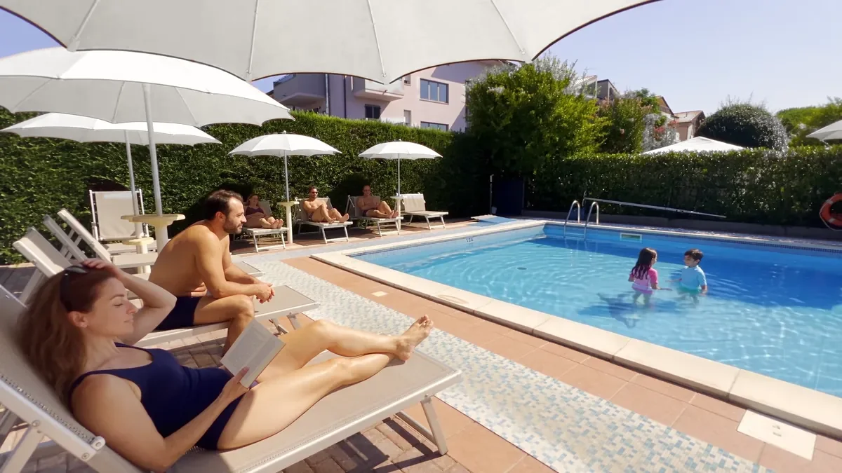 I genitori sui lettini del solarium sotto gli ombrelloni e i bambini che giocano in piscina